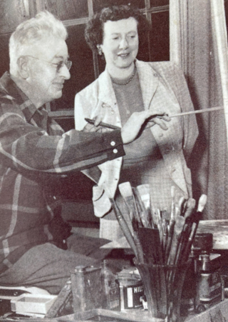 Sam painting at his easel in his home studio with his wife Marion looking on.